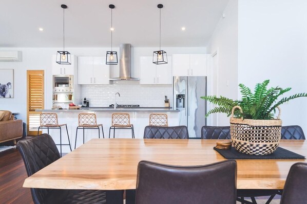 A modern kitchen with white cabinets, a wooden dining table, and hanging pendant lights. - The Salt House - Poolside amongst the Moonah Trees (Rye)