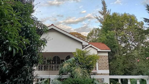 A two-story house with a red-tiled roof, a balcony with a white railing, and a garden with various plants and trees. - Stay.Plus karen Party House (Nairobi)
