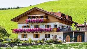 Une maison en bois à deux étages, dotée de balcons, avec un mur de pierre à l’avant et une colline en arrière-plan.