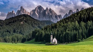 Eine kleine Kirche mit einem Glockenturm, umgeben von einem üppigen grünen Feld und einem bewaldeten Gebirgszug.