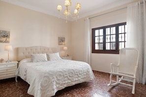 Une chambre à coucher avec un grand lit, un fauteuil à bascule blanc, un lustre et une fenêtre avec des rideaux.