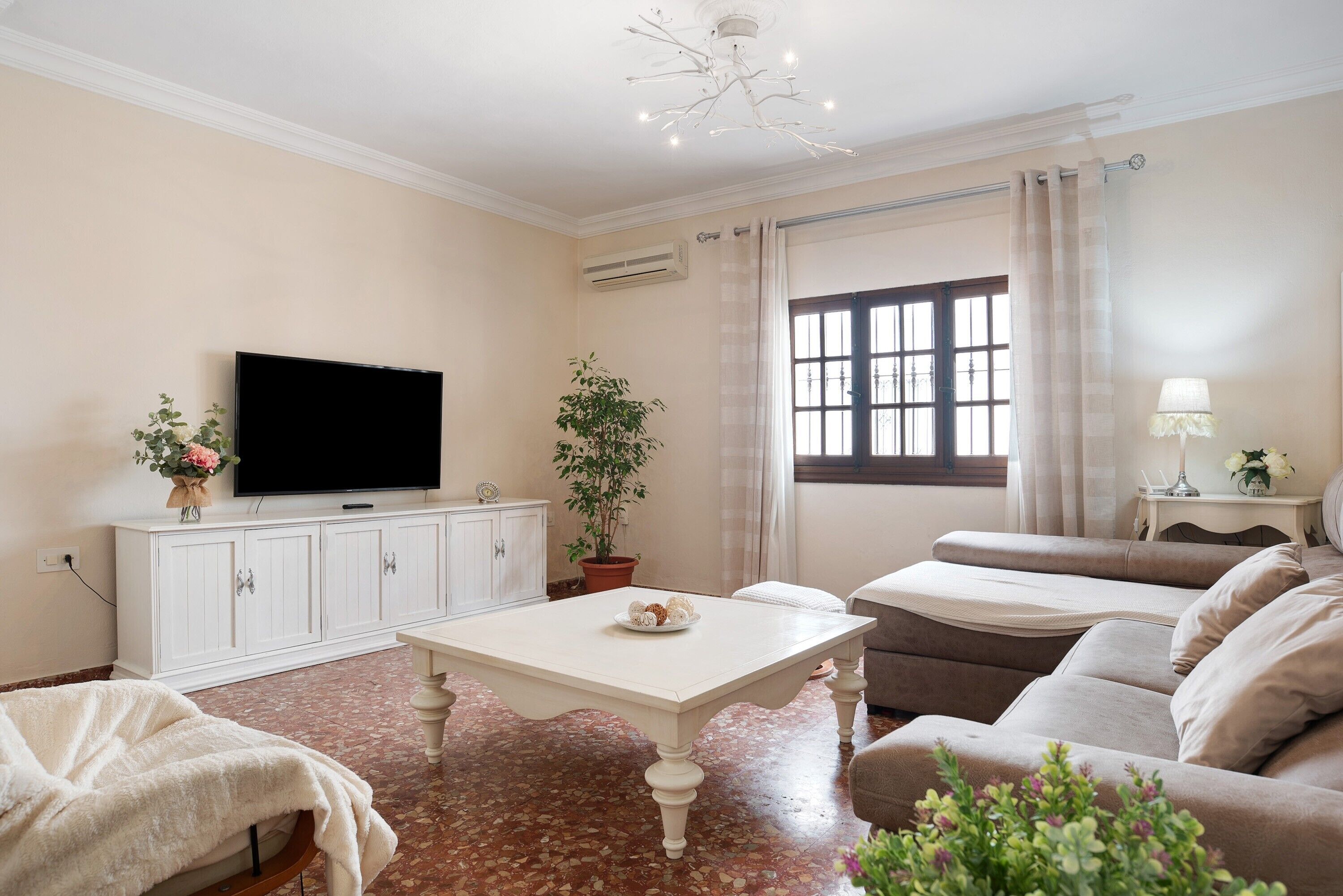 A living room with a flat-screen TV, a white cabinet, a potted plant, a beige sofa, and a window with curtains.