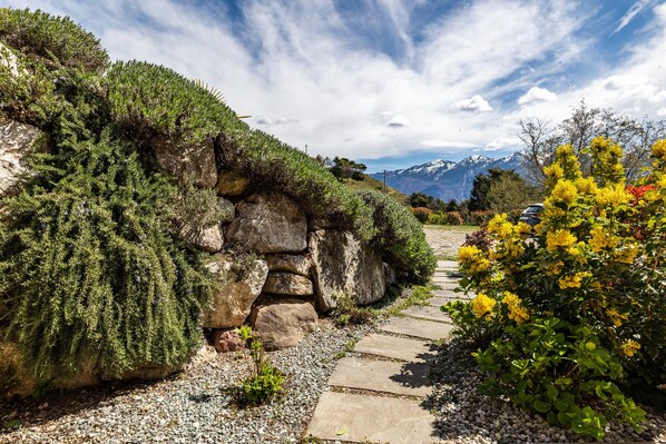 Ein Steinweg schlängelt sich durch einen Garten mit verschiedenen Pflanzen und einem Bergpanorama im Hintergrund.