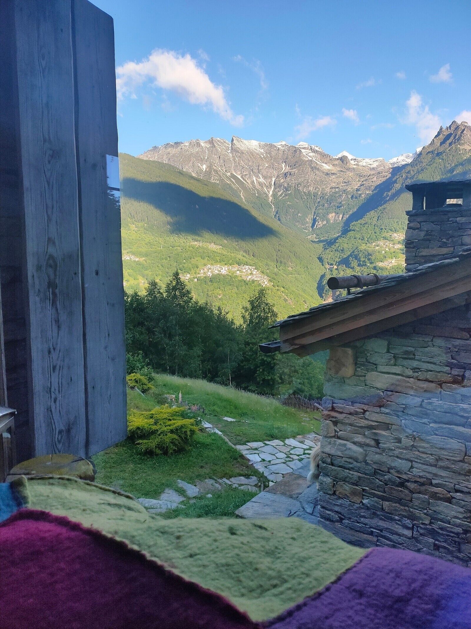 Ein rustikales Steinhaus mit Steinziegeldach, eine Holzkonstruktion links und eine gebirgige Landschaft im Hintergrund.