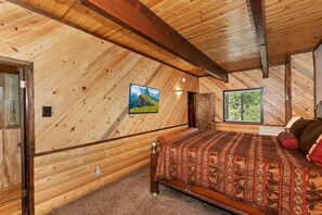 A bedroom with a wooden ceiling, a bed with patterned bedding, a mounted TV, and a window with a view of trees.