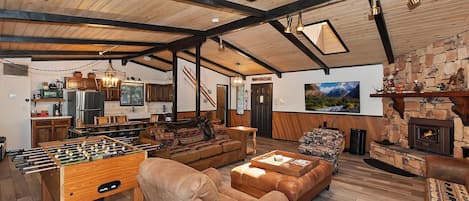 A cozy living room with a foosball table, leather sofas, a fireplace, and a kitchen area with wooden cabinets and a hanging light fixture.