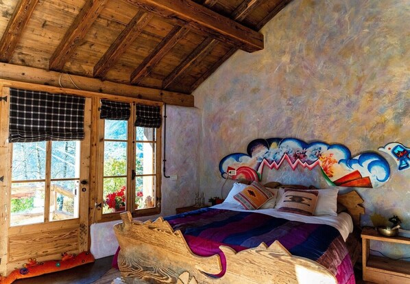 Ein rustikales Schlafzimmer mit einem Holzkopfende, einem Bett mit bunter Bettwäsche, einem Fenster mit karierter Gardine und einer Wand mit Wandmalerei.