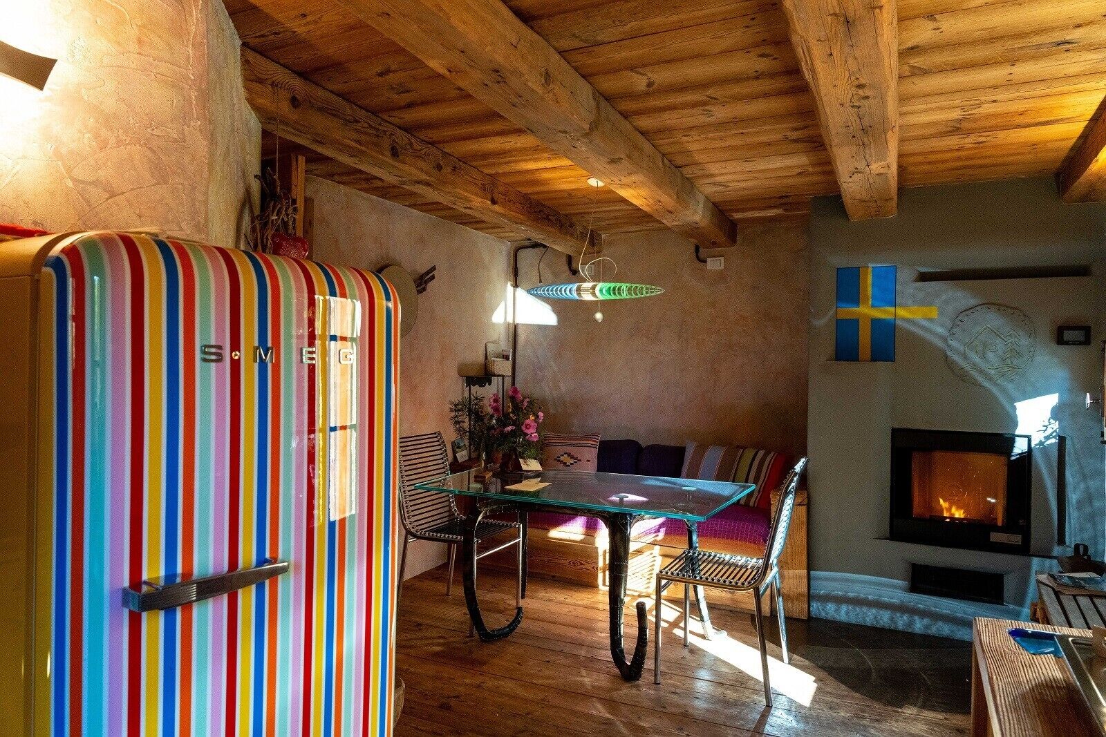 A cozy room with a striped refrigerator, a glass-top dining table, wooden chairs, a sofa, and a fireplace.