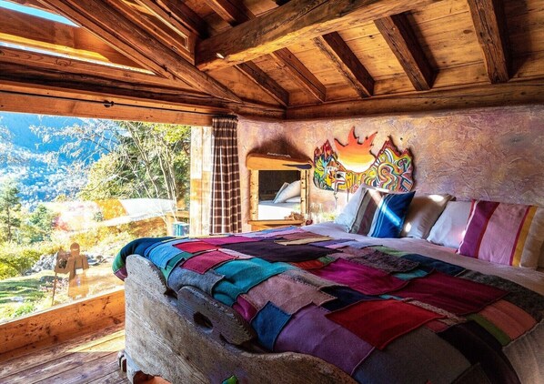 Une pièce rustique en bois, avec un lit, une fenêtre donnant sur un paysage montagneux et une fresque colorée sur le mur.
