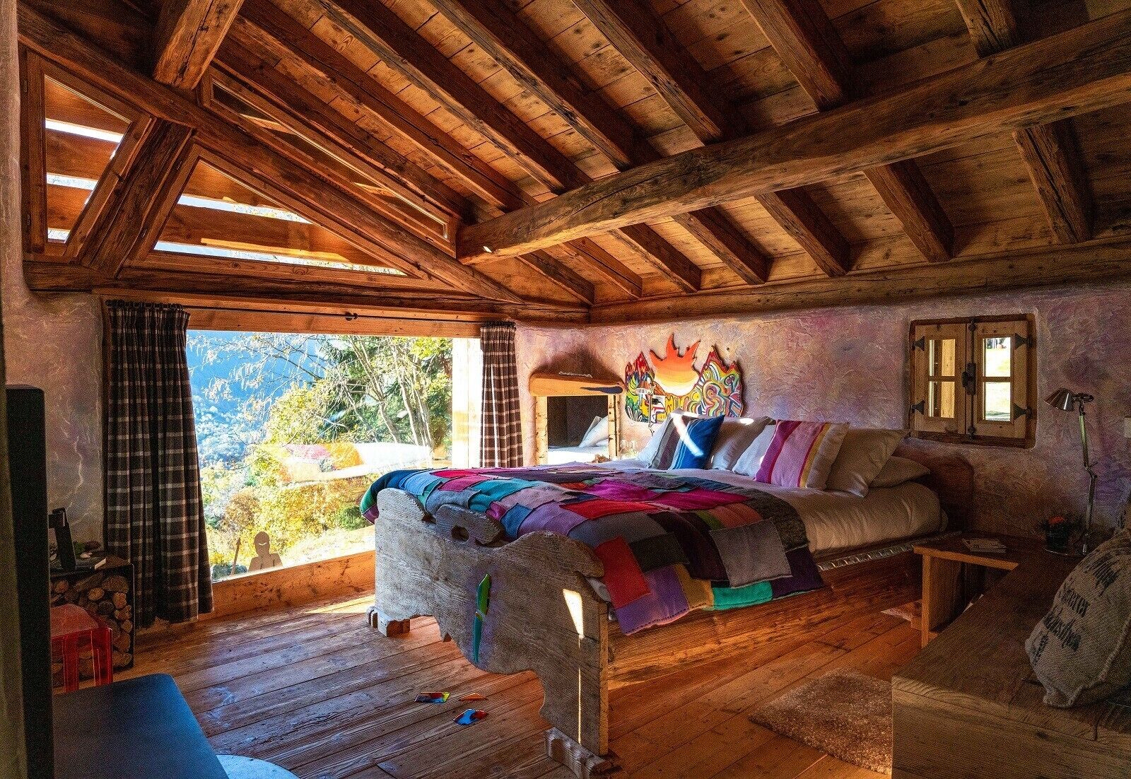 A rustic wooden room with a bed, a sofa, and a window overlooking a mountainous landscape.