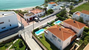 Et kystnære boligområde med hvide bygninger, en swimmingpool og en strand i baggrunden.