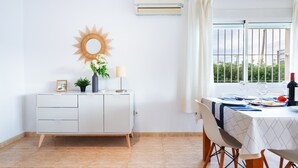 Et spiseområde med et bord dækket til to, et hvidt skab med skuffer, et solformet spejl og en aircondition.