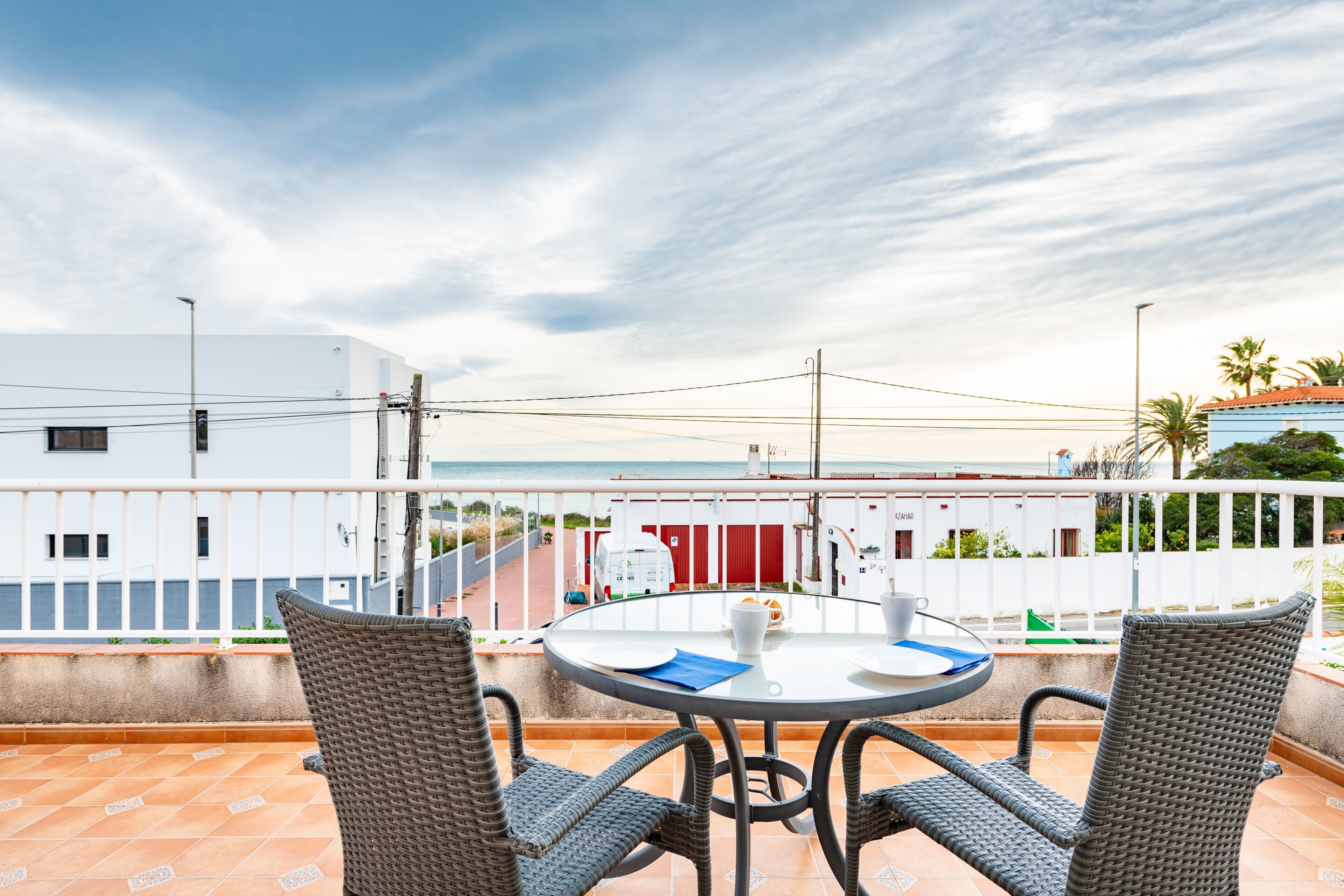 Un balcón con sillas de mimbre, una mesa redonda y vistas al mar y edificios.