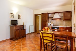 A kitchen with wooden cabinets, a dining table with chairs, a fruit bowl, and a television on a cabinet. - Agriturismo 'Bilo Erica' with Pool, Wi-Fi & Garden (Marina di Grosseto)
