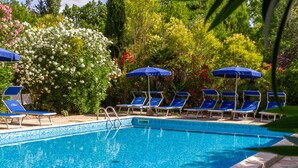 A swimming pool with blue lounge chairs and umbrellas, surrounded by lush greenery and flowering plants.
