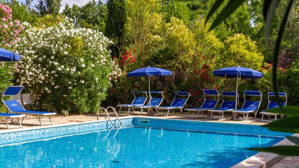 Ein Swimmingpool mit blauen Liegestühlen und Sonnenschirmen, umgeben von üppiger Vegetation und blühenden Pflanzen.