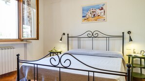 Un dormitorio con cama, ventana con cortinas, un cuadro en la pared y una mesita de noche con lámpara.