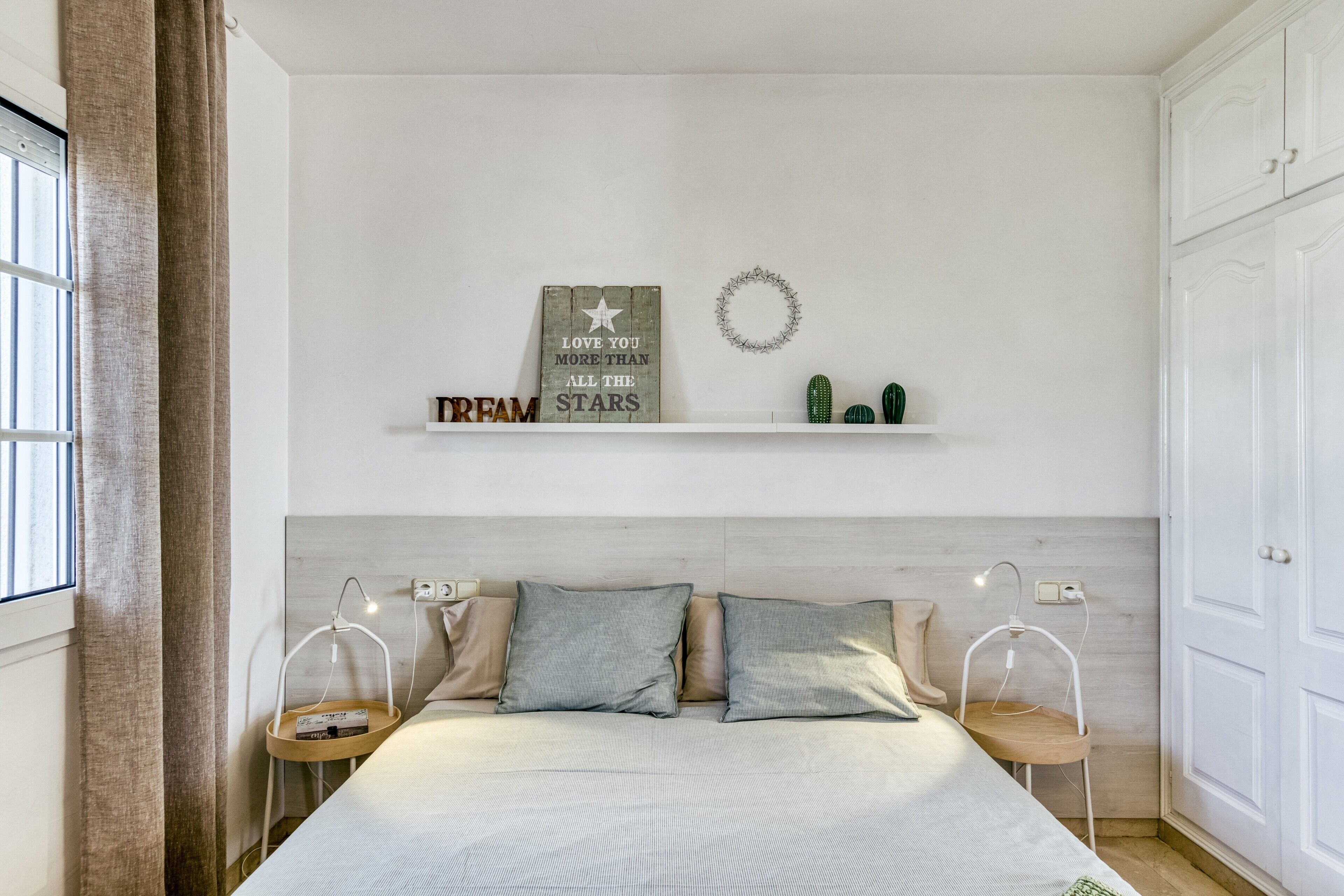 Ein Schlafzimmer mit einem Bett, Nachttischen, einem an der Wand befestigten Regal mit Dekoration und einem Fenster mit Vorhängen.