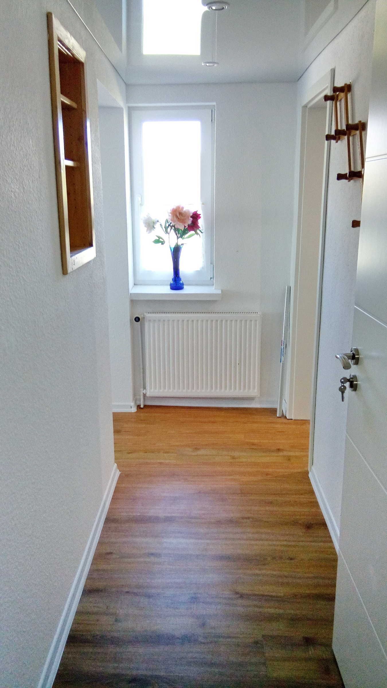 A hallway with a wooden floor, a white radiator, a window with a vase of flowers, and a door with a key lock.