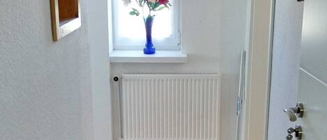A hallway with a wooden floor, a white radiator, a window with a vase of flowers, and a door with a key lock.