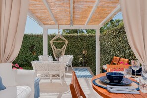 Un espacio exterior techado con zona de estar, una mesa dispuesta para comer, muebles de mimbre blanco y una silla colgante.