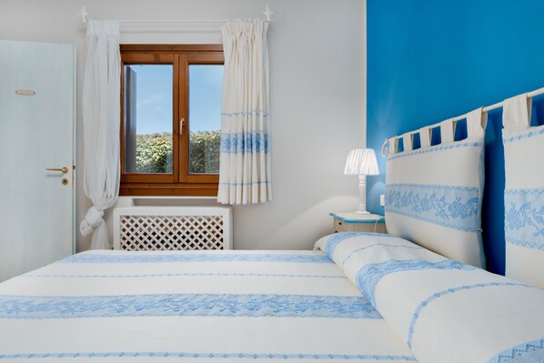 Un dormitorio con cama, ventana con cortinas, radiador, mesita de noche con lámpara y pared con detalle en azul.