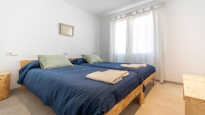 A bedroom with two beds, a window with curtains, and a small bedside table. - Apartment 'Guillem' with Sea Views, Garden, and Wi-Fi (Cala Sant Vicenç)