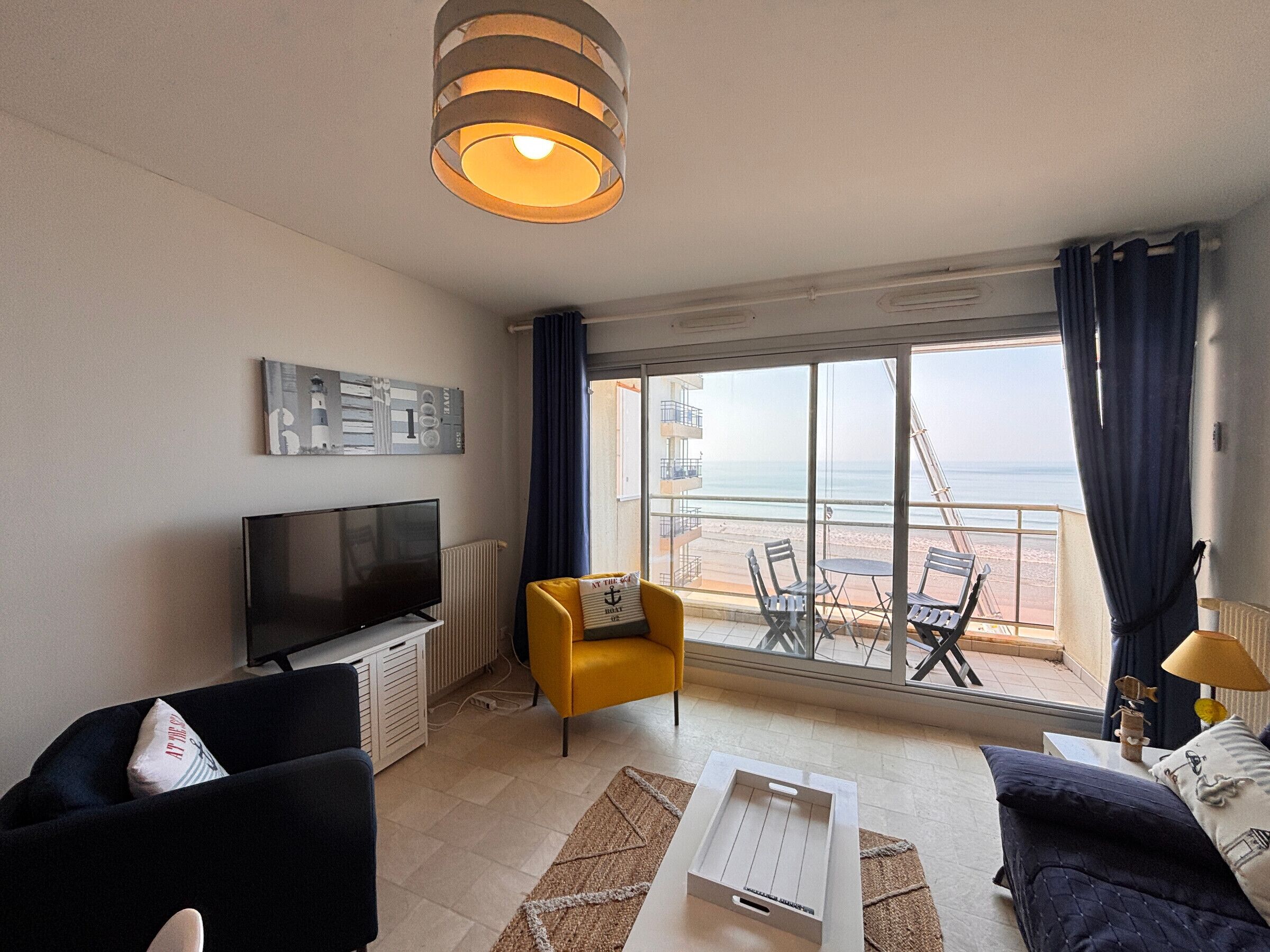 Una sala de estar moderna con televisor, sofá, sillón y vistas a la playa a través de amplias ventanas.