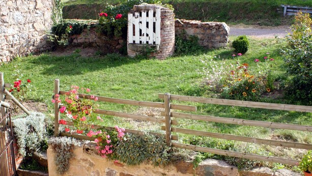 Un mur de pierre avec une porte en bois, un bâtiment en pierre et une zone herbeuse parsemée de fleurs.