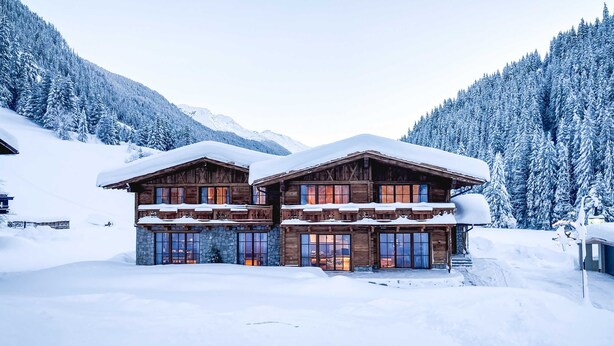 Un chalet en bois, doté de fondations en pierre, de plusieurs balcons et offrant une vue sur des montagnes enneigées.