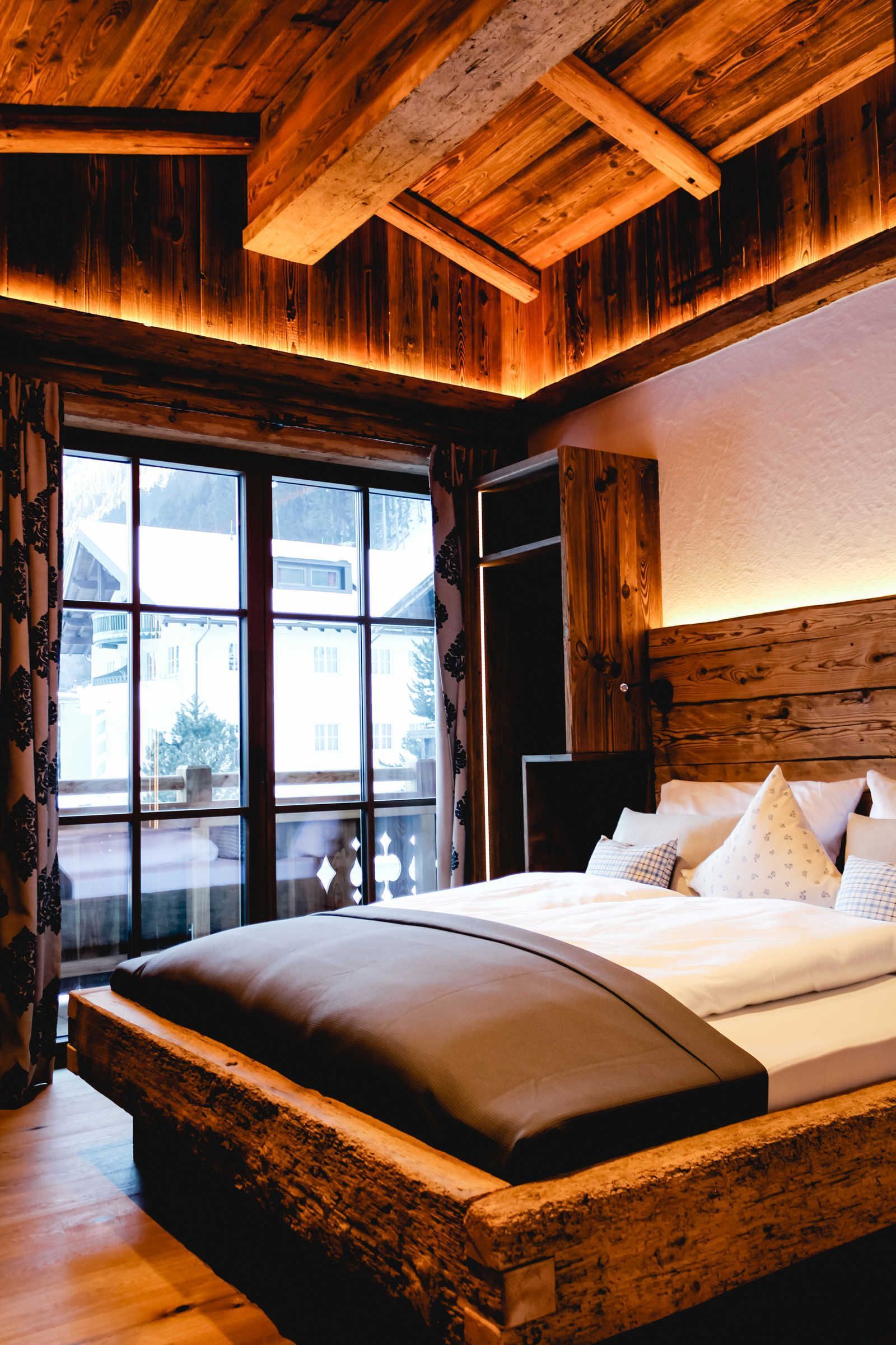 Een slaapkamer met houten frame, een groot raam, een bed met witte beddengoed en een houten hoofdbord.