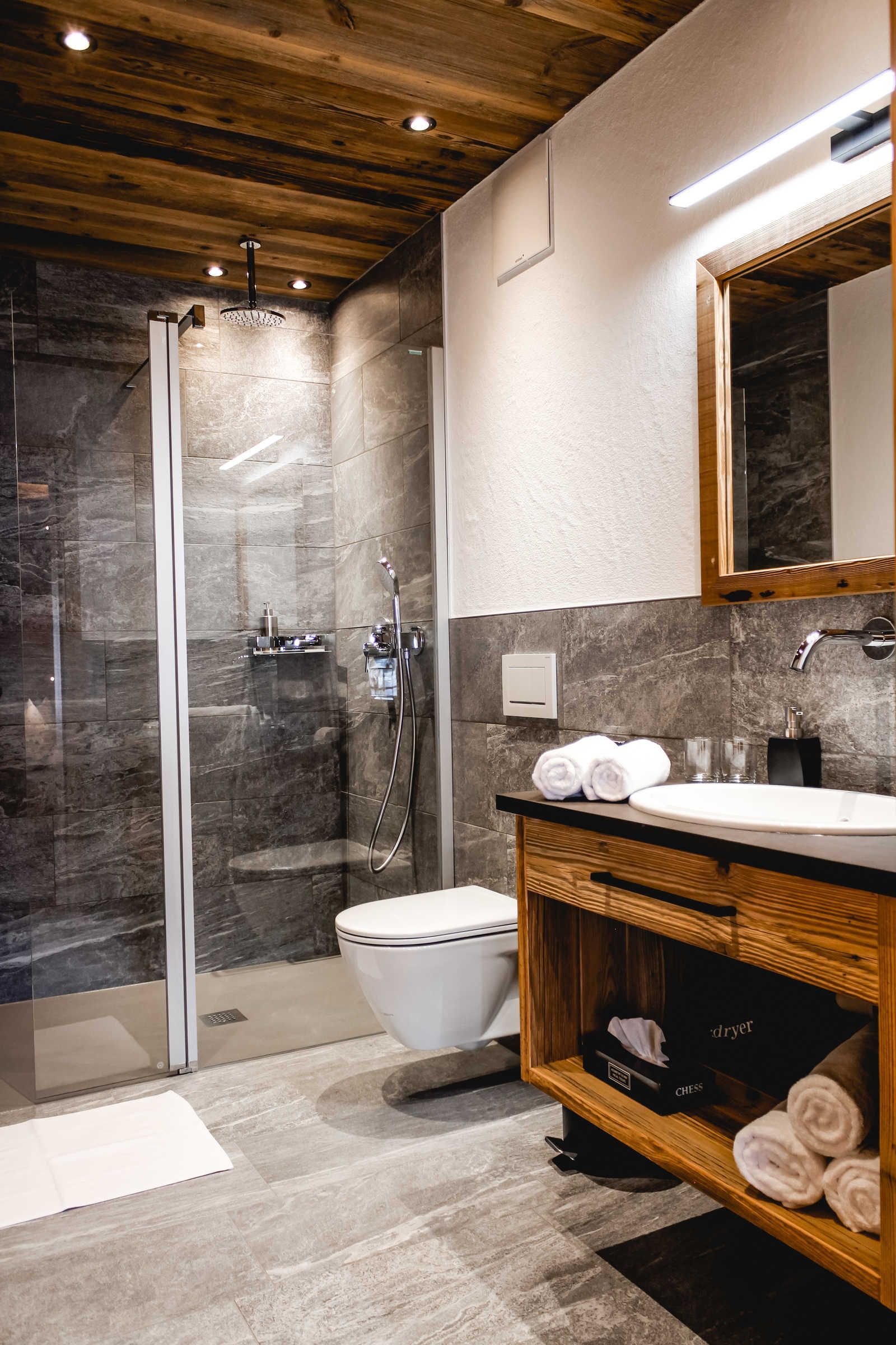 A modern bathroom with a glass-enclosed shower, a white toilet, a wooden vanity with a mirror, and neatly rolled towels.