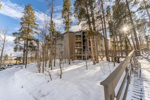 A building surrounded by snow-covered ground and trees, with a wooden deck in the foreground. - Treehouse #301, Building A by Summit County Mountain Retreats (Silverthorne)