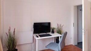 A room with a white desk, a flat-screen TV, a grey chair, and two potted plants. - Apartment city district 1 (Bremerhaven)