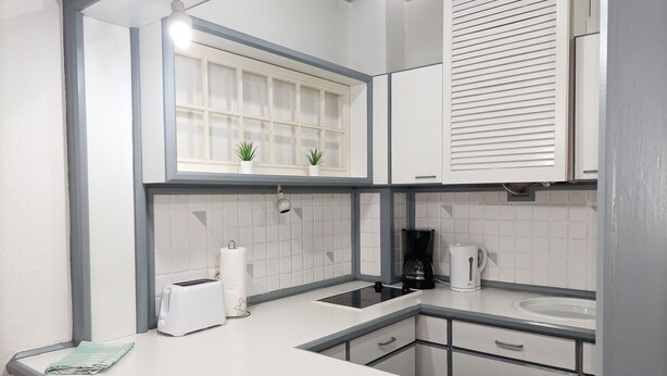 A modern kitchen with white cabinets, a black coffee maker, a white kettle, and a window with plants. - Apartment city district 1 (Bremerhaven)