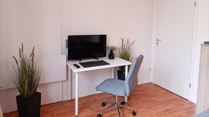 A small, tidy room with a white desk, a grey office chair, a flat-screen TV, and two potted plants. - Apartment city district 1 (Bremerhaven)