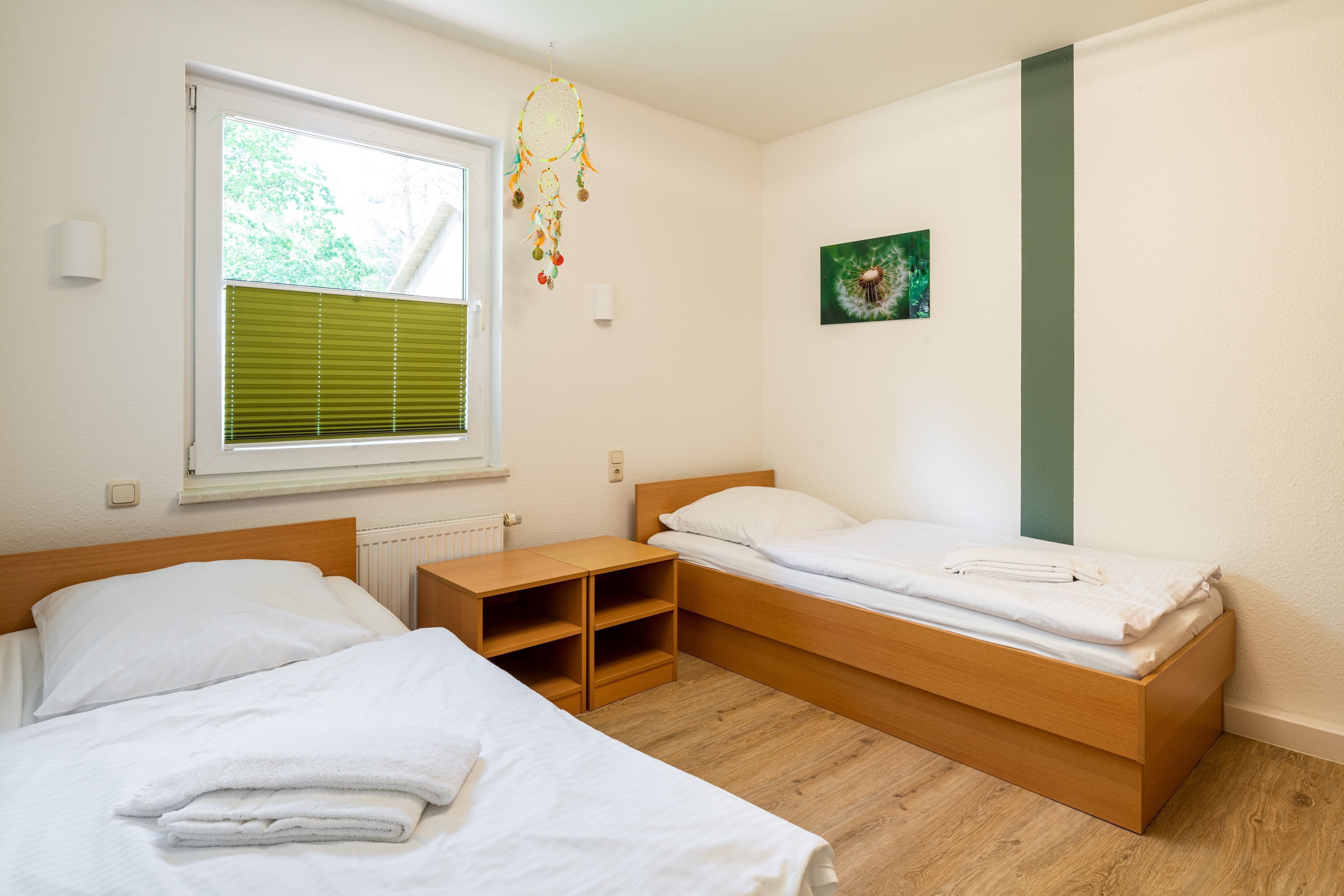 Un dormitorio con dos camas, una ventana con persianas, una mesita de noche y un atrapasueños colgado en la pared.