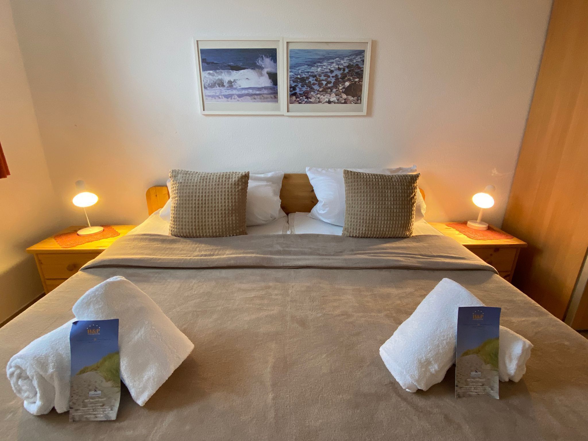 Een net opgemaakt bed met twee kussens, een deken en twee handdoeken met brochures erop. Twee nachtkastjes met lampen en een muur met twee ingelijste foto's.