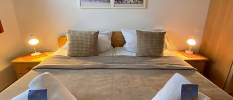 Una cama bien hecha con dos almohadas, una colcha y dos toallas con folletos encima. Hay dos mesitas de noche con lámparas y una pared con dos cuadros enmarcados.