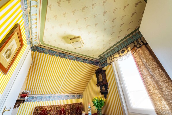 A room with striped wallpaper, a patterned ceiling, a wooden clock, and a window with curtains.