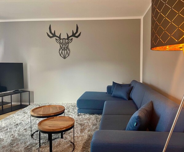 A living room with a sectional sofa, two wooden coffee tables, a mounted deer head wall art, and a TV on a stand.
