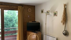Ett rum med ett träpanelat tak, ett rutigt bord, en tv på ett träskåp, ett element och ett fönster med gardiner.
