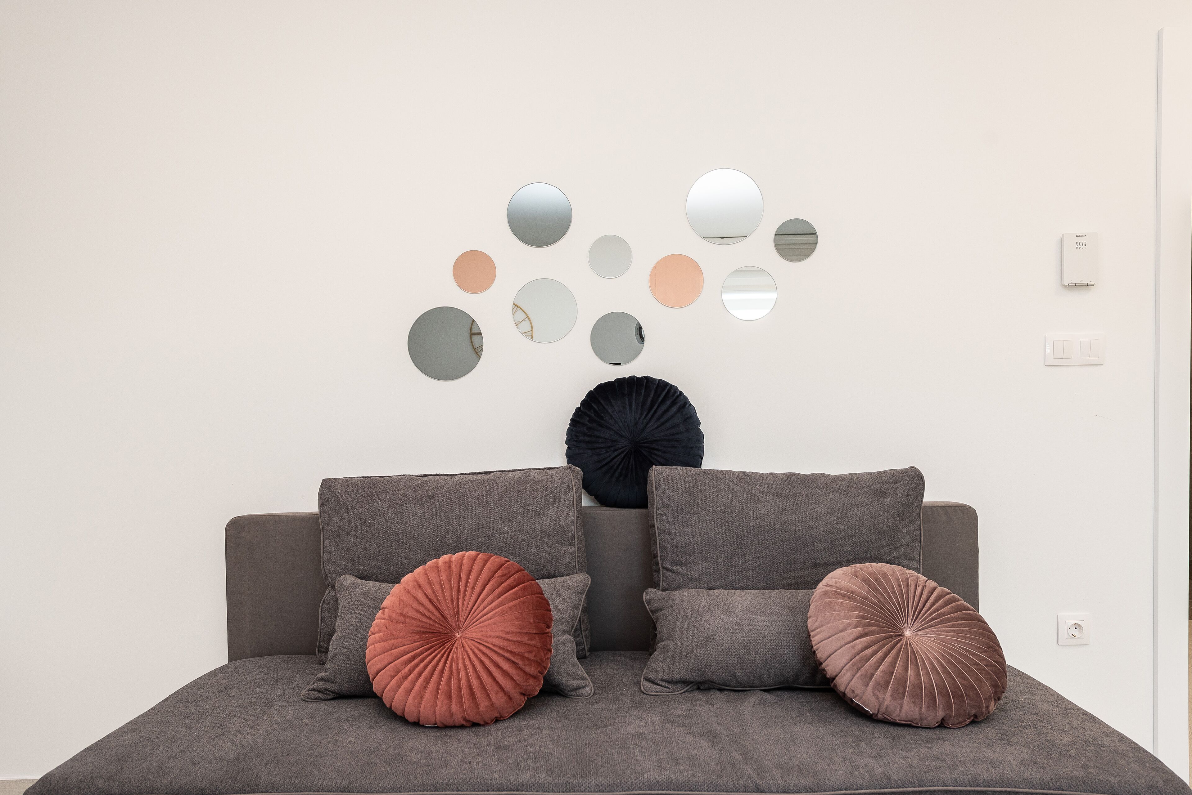 A modern living room with a grey sofa, two large round cushions, and a wall-mounted mirror arrangement.