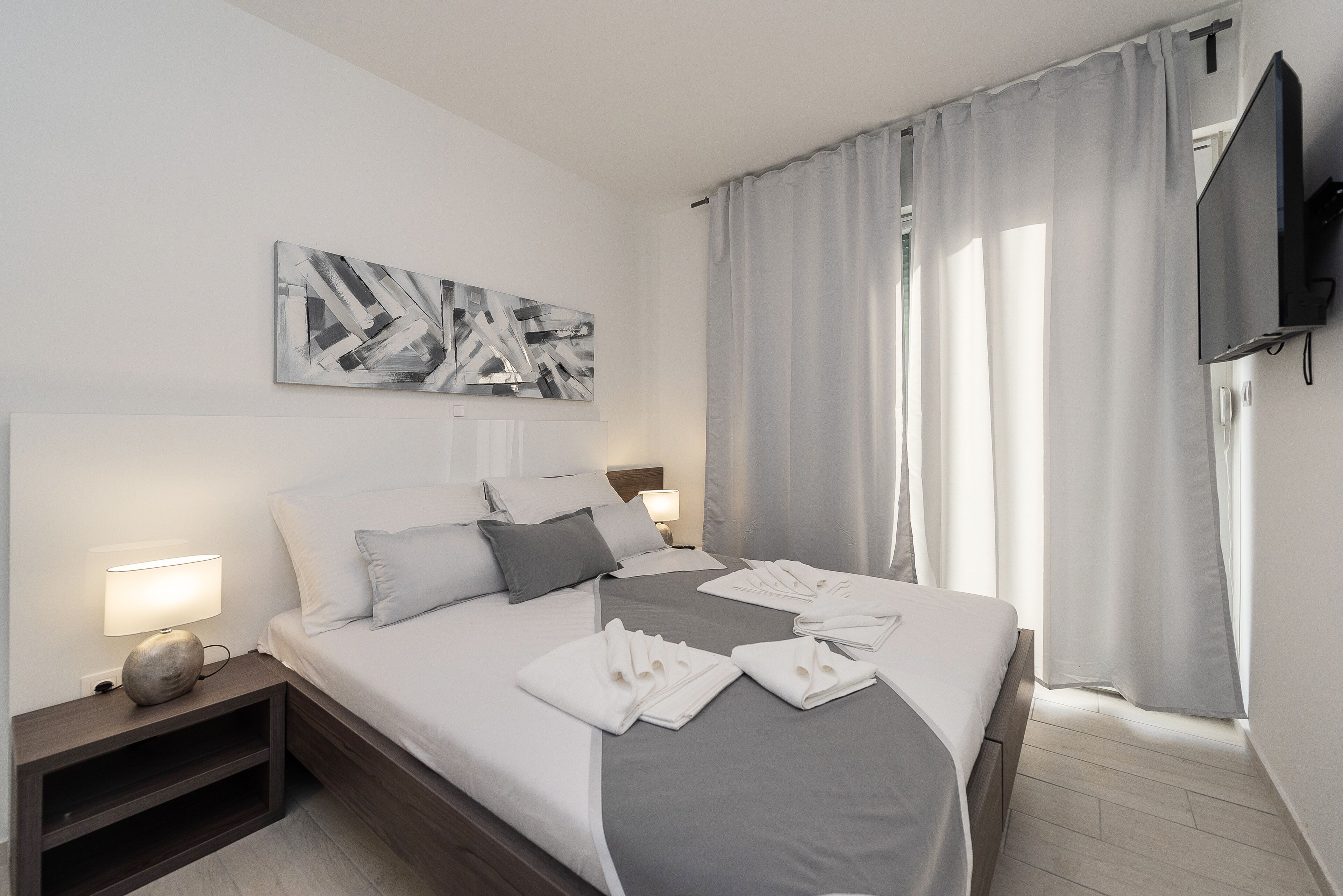 A modern hotel room with a neatly made bed, a flat-screen TV mounted on the wall, and a large window with sheer curtains.