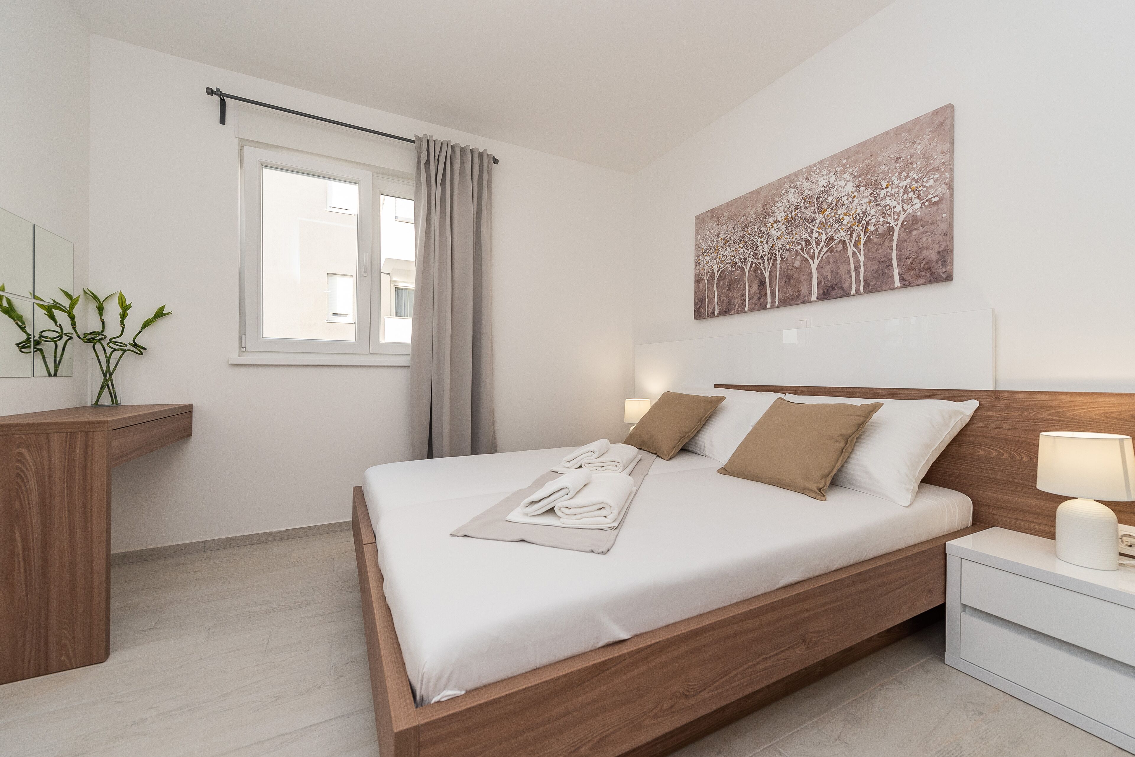 Un dormitorio ordenado con una cama de madera, ropa de cama blanca, una mesita de noche con una lámpara, una ventana con cortinas y un cuadro colgado en la pared.