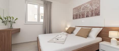 Un dormitorio ordenado con una cama de madera, ropa de cama blanca, una mesita de noche con una lámpara, una ventana con cortinas y un cuadro colgado en la pared.
