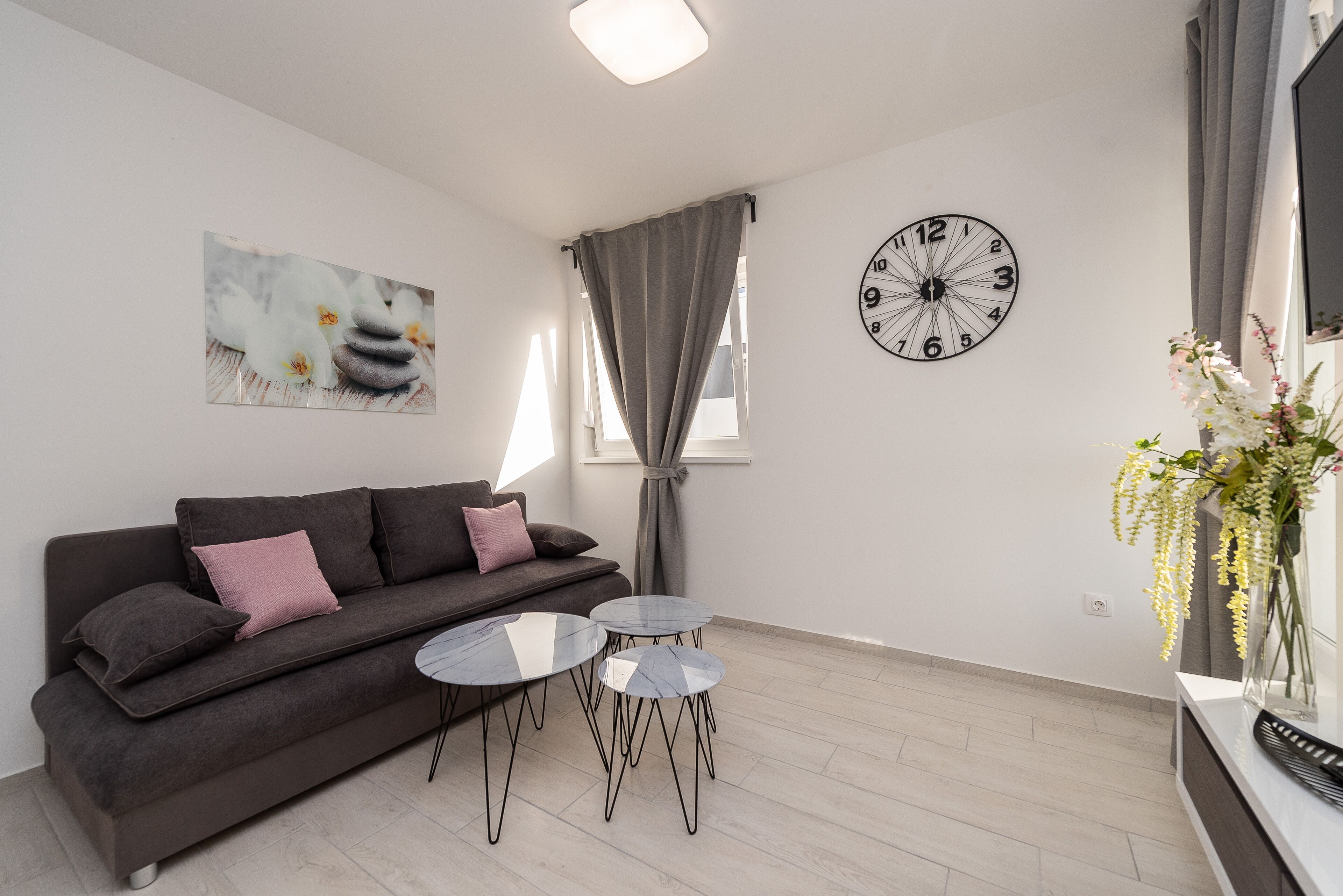 A modern living room with a dark grey sofa, two small round tables, a wall clock, and a painting of flowers.