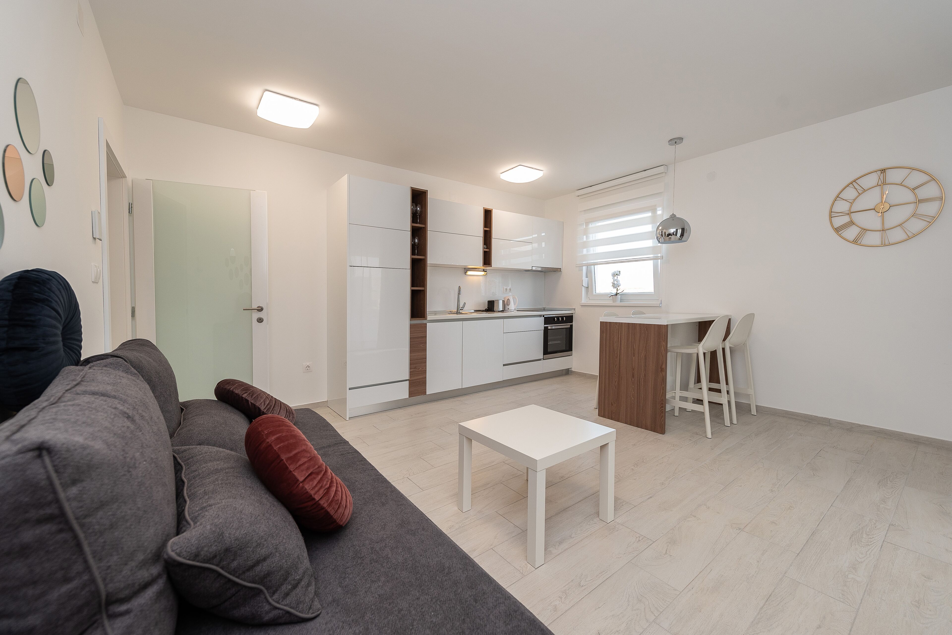 A modern living room with a grey sofa, a white coffee table, and a kitchen area with white cabinets and a wooden countertop.