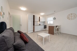 Una sala de estar moderna con un sofá gris, una mesa de centro blanca y una zona de cocina con armarios blancos y encimera de madera.