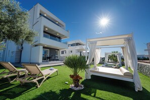Un espacio lounge exterior moderno con una tumbona y sillones, un césped cuidado y un edificio grande al fondo.